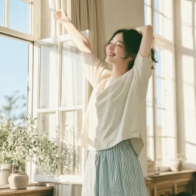 朝の光が差しこむ部屋で、窓辺に立って両腕を上げて伸びをする女性