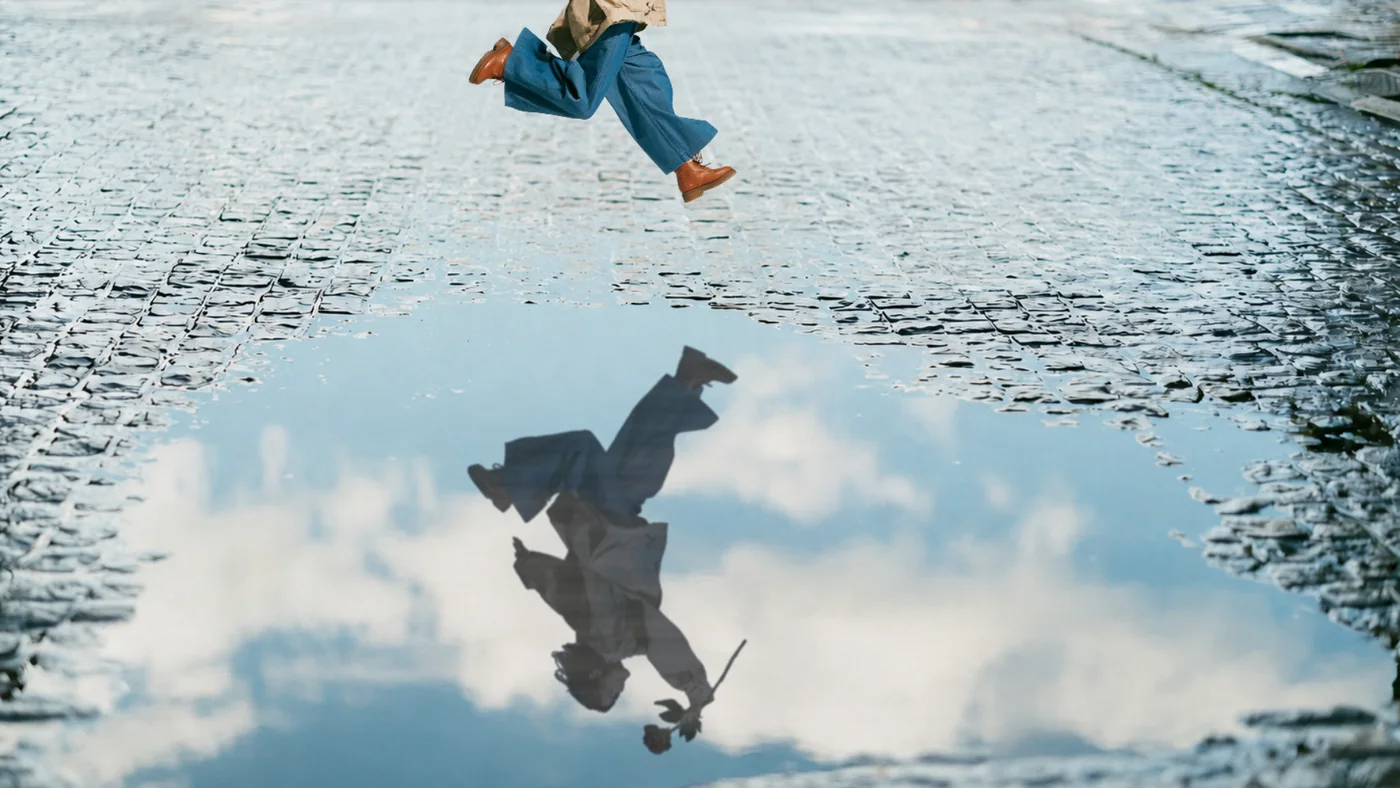 雨上がりの石畳。水たまりを飛び越える女性の脚と、水面に映る青空とその全身